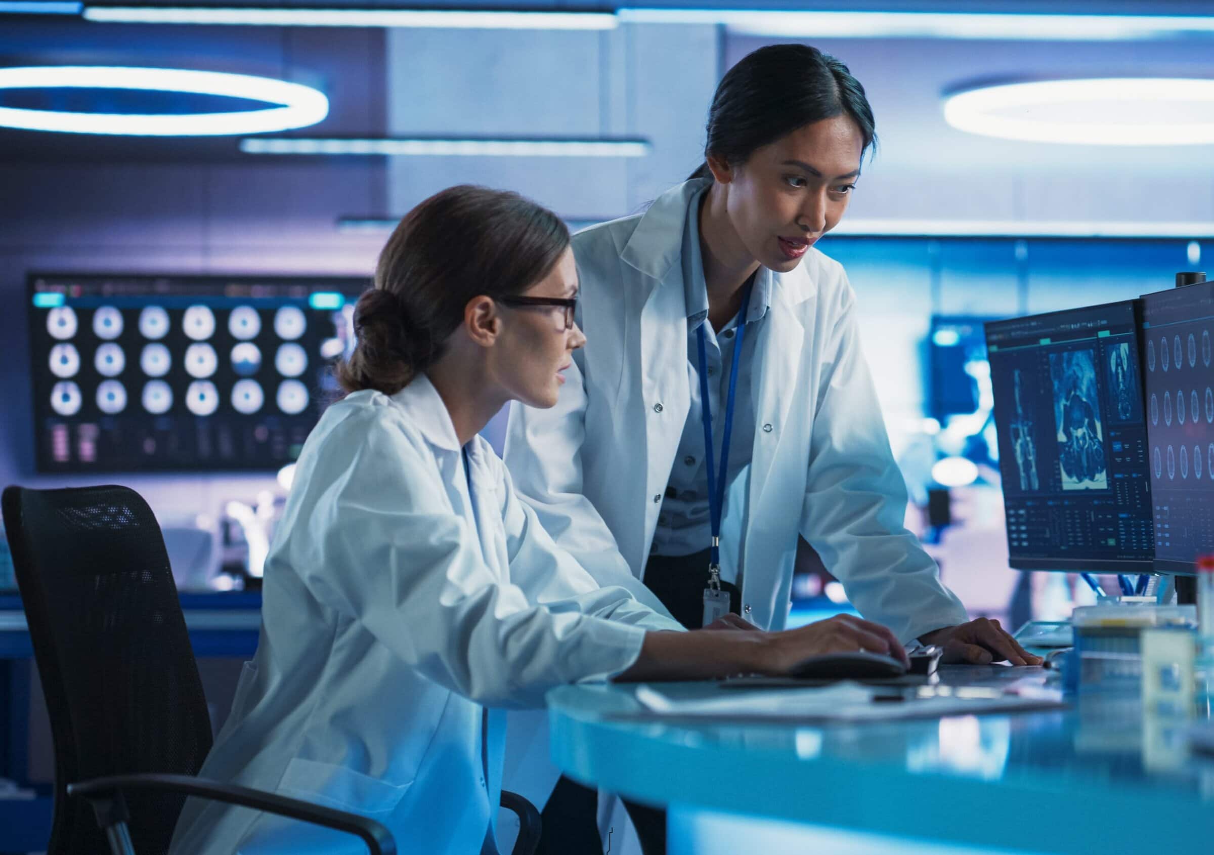 Asian And Caucasian Female Doctors Talking In Medical Research Center And Using Desktop Computer To Analyze MRI Scans Of Brain. Surgeon And Neuroscientist Discussing Solutions For Treating Alzheimer's